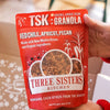Red package of TSK Granola held by a person with a blurred indoor background