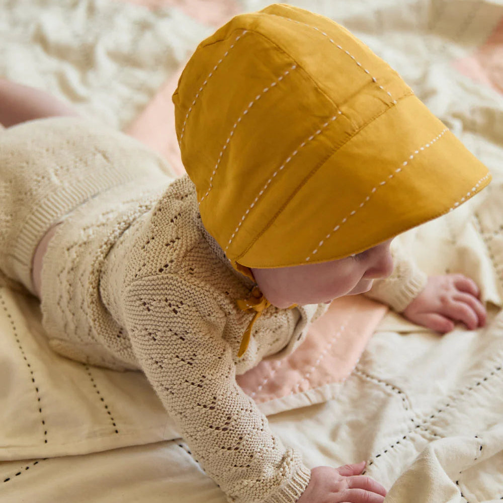 Baby wearing a yellow bonnet and beige knitted outfit on a soft surface