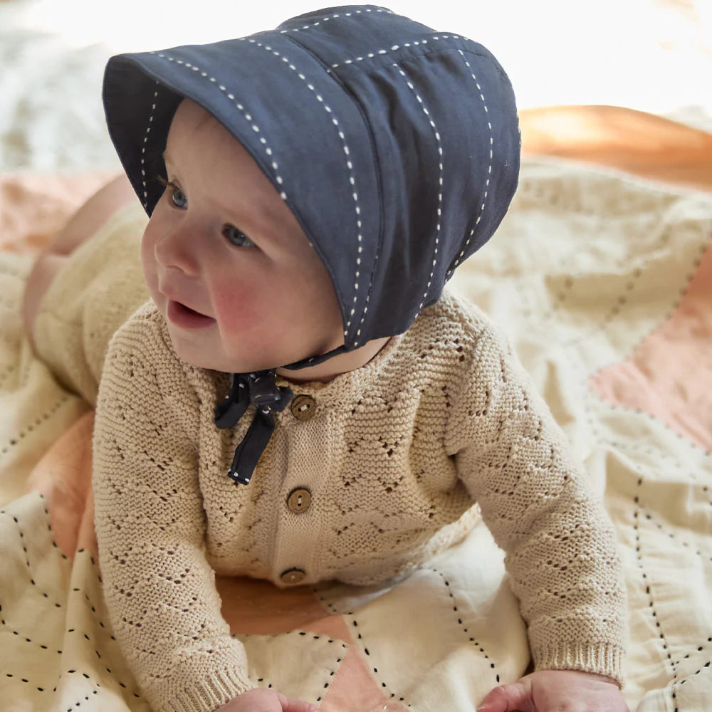 Baby wearing a navy blue bonnet and beige knitted sweater on a soft surface