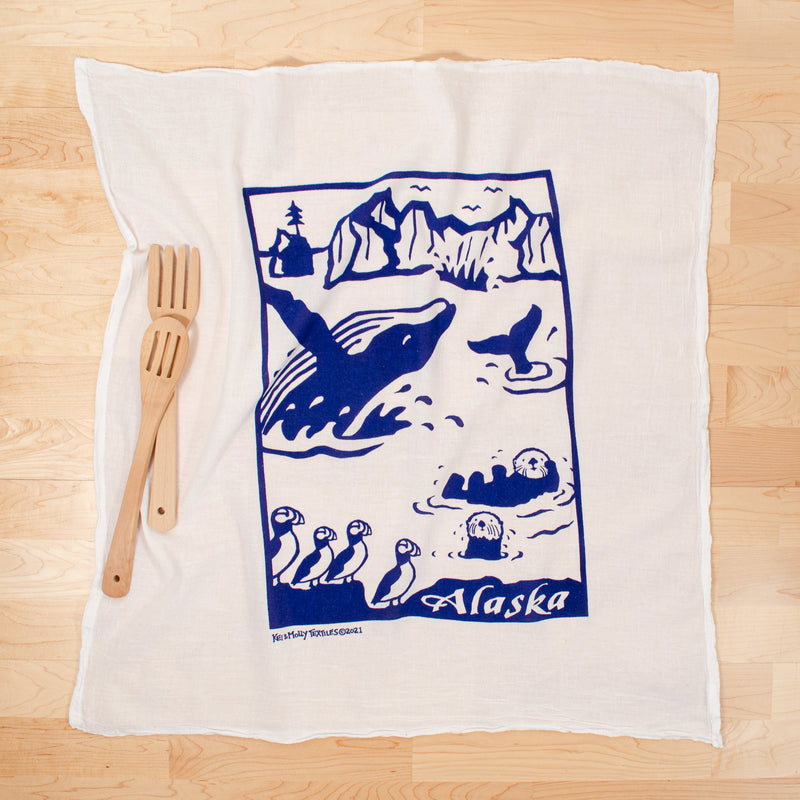 Kei & Molly illustration: Whale swimming in Alaska surrounded by otters and puffins printed in Navy Blue on white cotton flour sack dish towel propped with two wooden spoons