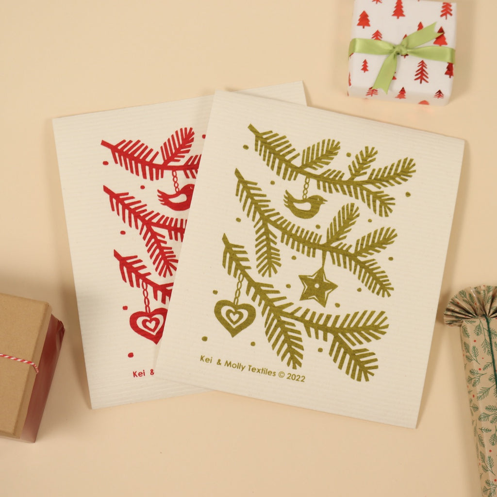 Two Kei & Molly Textiles Large Sponge Cloths “Pine branches!” design printed in Green and Red propped with presents. 