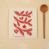 Kei & Molly Textiles Large Sponge Cloth “Pine branches!” design printed in red propped with wooden spoon and snowflakes.