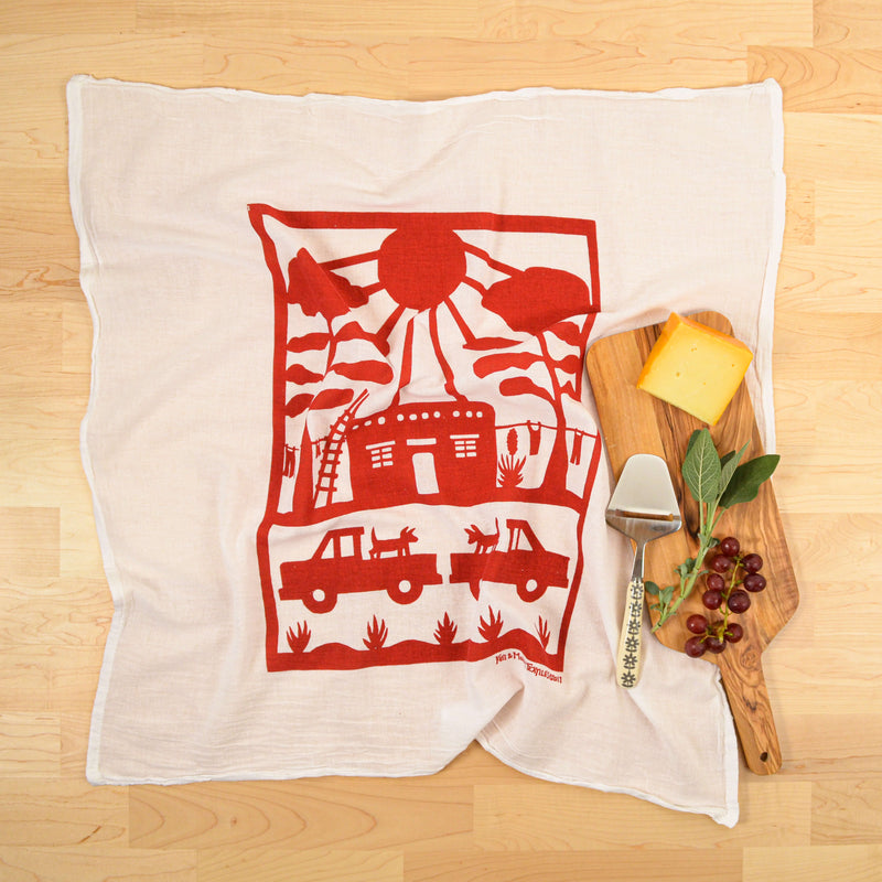 Kei & Molly Flour Sack Dish Towel: Adobe house with trucks, dogs and laundry line printed in color Red propped with cutting board with cheese and grapes